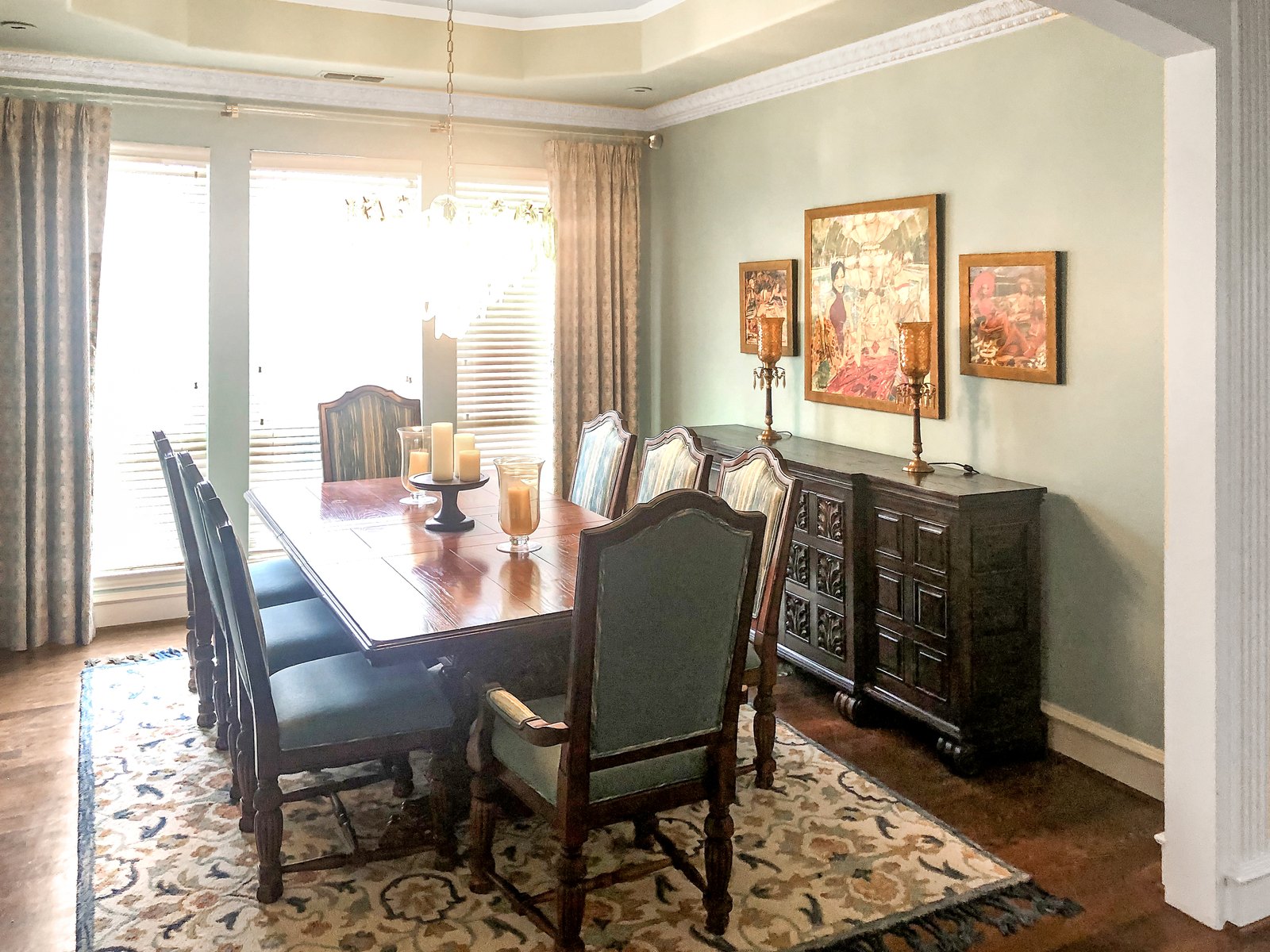 Sage green dining room with a dark wood table, blue upholstered chairs, a dark buffet and three framed figurative paintings