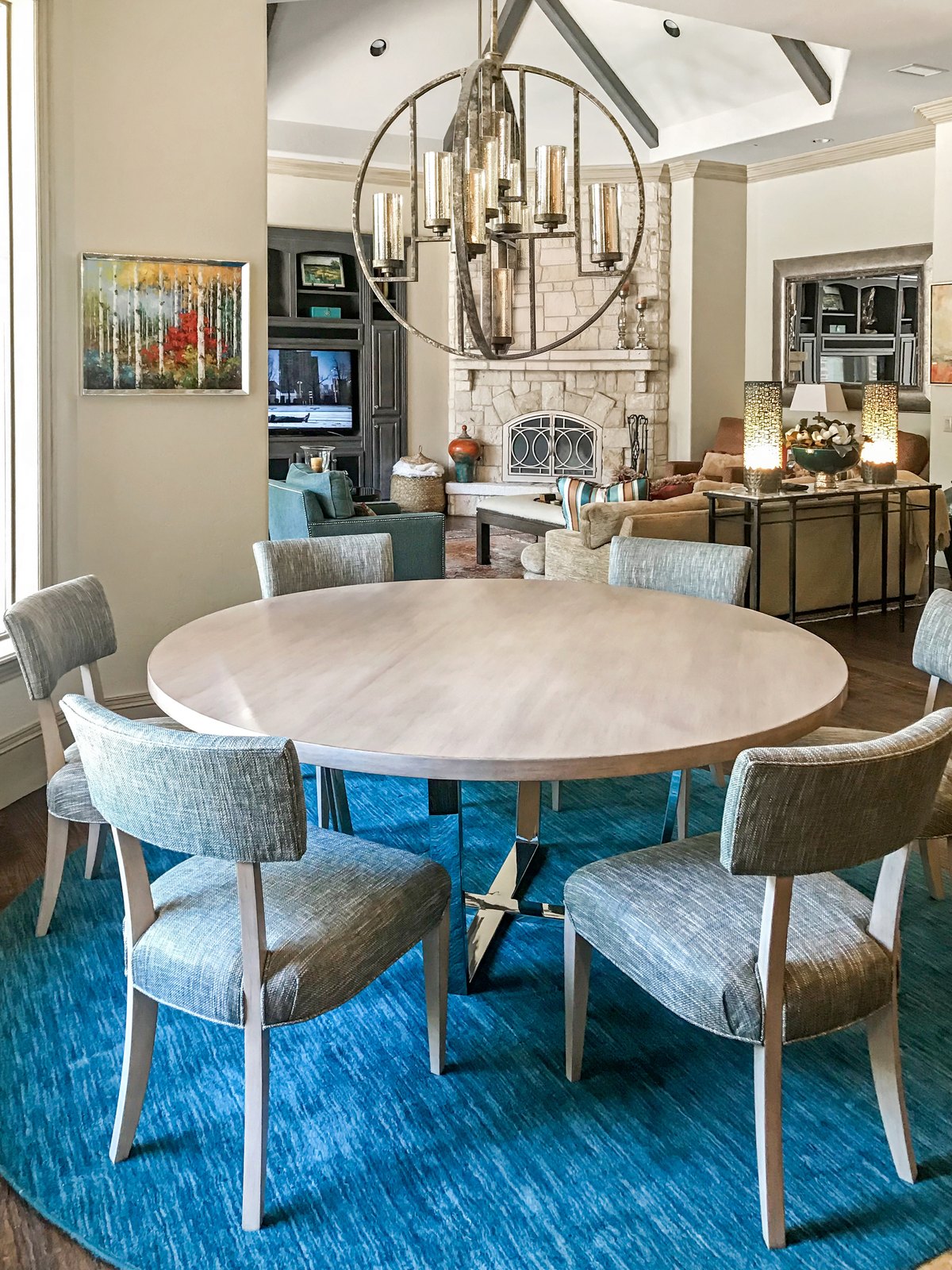Round breakfast table with upholstered chairs on a teal rug beneath an open-cage chandelier